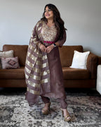 A woman models a brown ethnic suit set featuring a brown kurta with a gold sequined bodice, matching brown trousers, and a striped brown and gold dupatta, standing in a living room in front of a brown leather sofa.