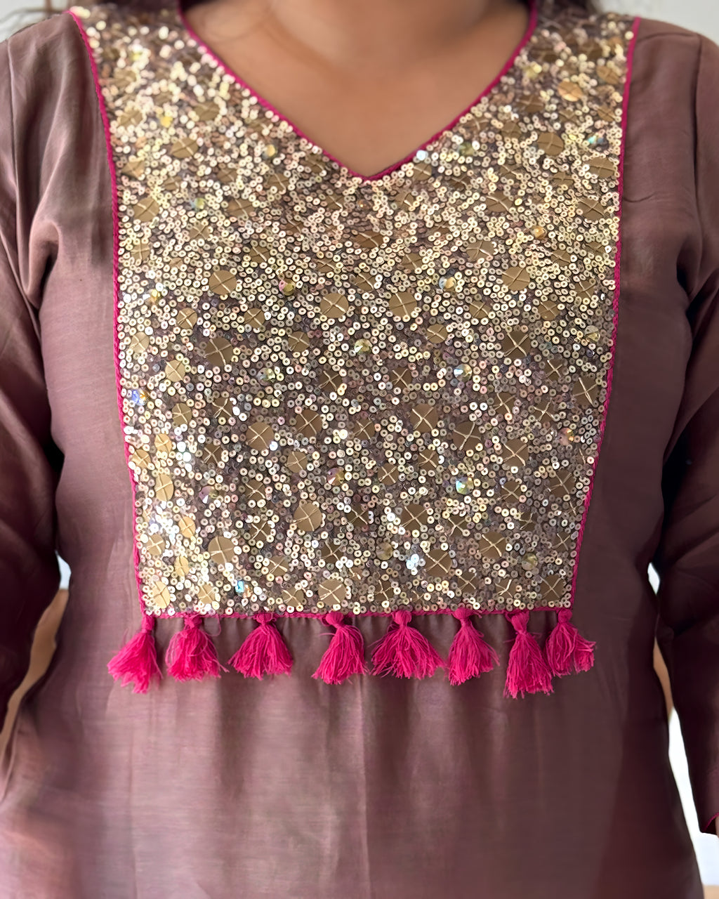 Close-up of a brown or mauve-colored traditional tunic top featuring a large, heavily embellished gold sequin and mirror work yoke panel with bright pink tassel trim along the neckline and hem.