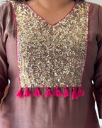 Close-up of a brown or mauve-colored traditional tunic top featuring a large, heavily embellished gold sequin and mirror work yoke panel with bright pink tassel trim along the neckline and hem.