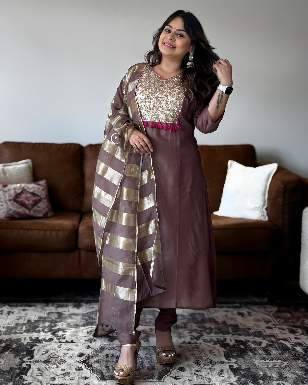 A woman models a brown Indian ethnic outfit, featuring a solid brown kurta with a heavily embellished gold sequined yoke, a matching brown dupatta with gold stripes, and brown trousers, set against a living room background with a brown sofa.