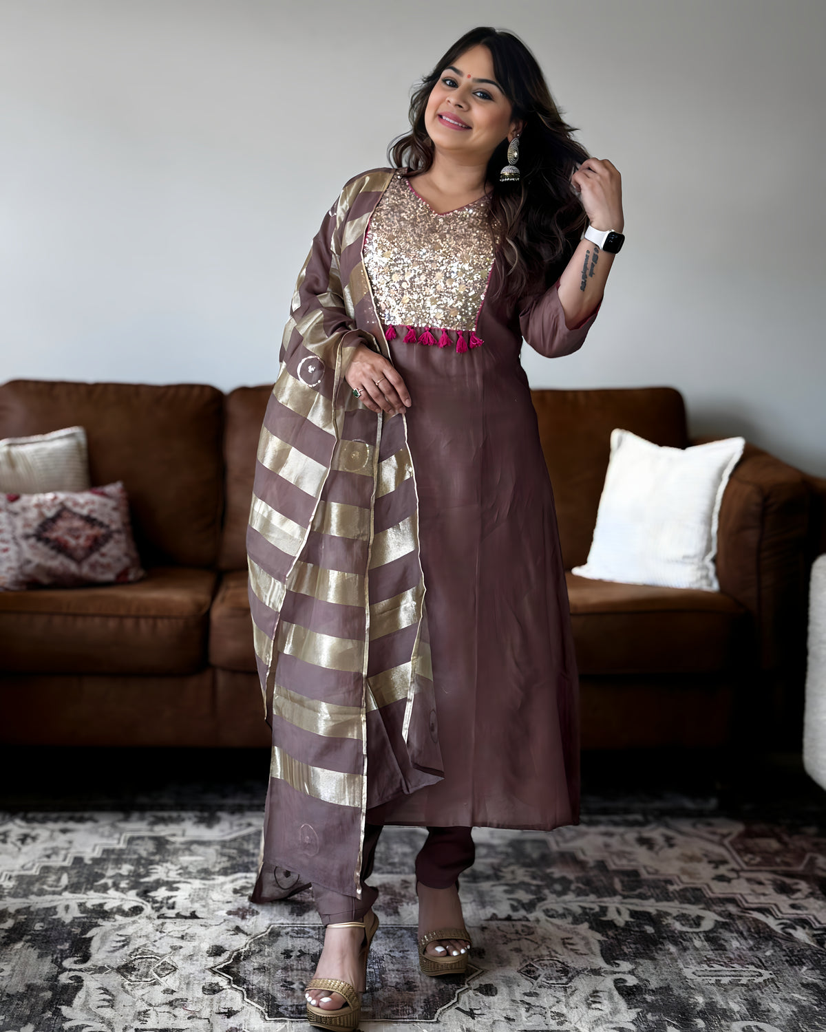 A woman models a brown Indian ethnic outfit, featuring a solid brown kurta with a heavily embellished gold sequined yoke, a matching brown dupatta with gold stripes, and brown trousers, set against a living room background with a brown sofa.