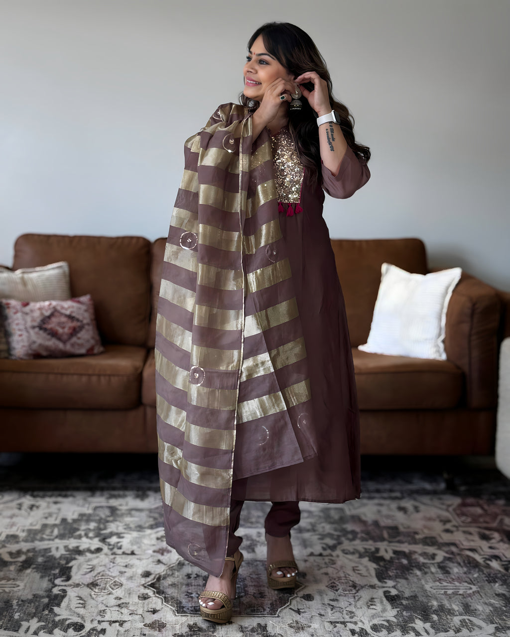 A woman models a traditional Indian outfit consisting of a brown kurta with gold sequin embroidery on the chest, matching brown trousers, and a brown and gold striped dupatta, standing in a living room with a brown sofa in the background.