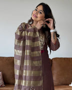 A woman wearing a brown and gold striped dupatta and a brown embroidered kurta, posing in front of a brown leather sofa.