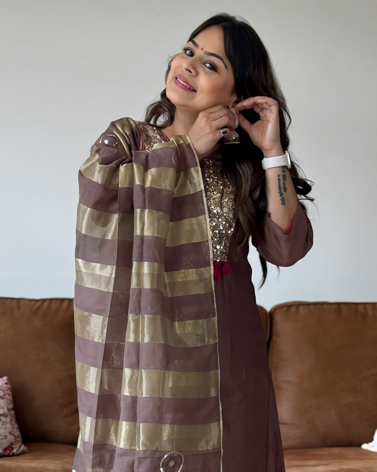 A woman wearing a brown and gold striped dupatta and a brown embroidered kurta, posing in front of a brown leather sofa.