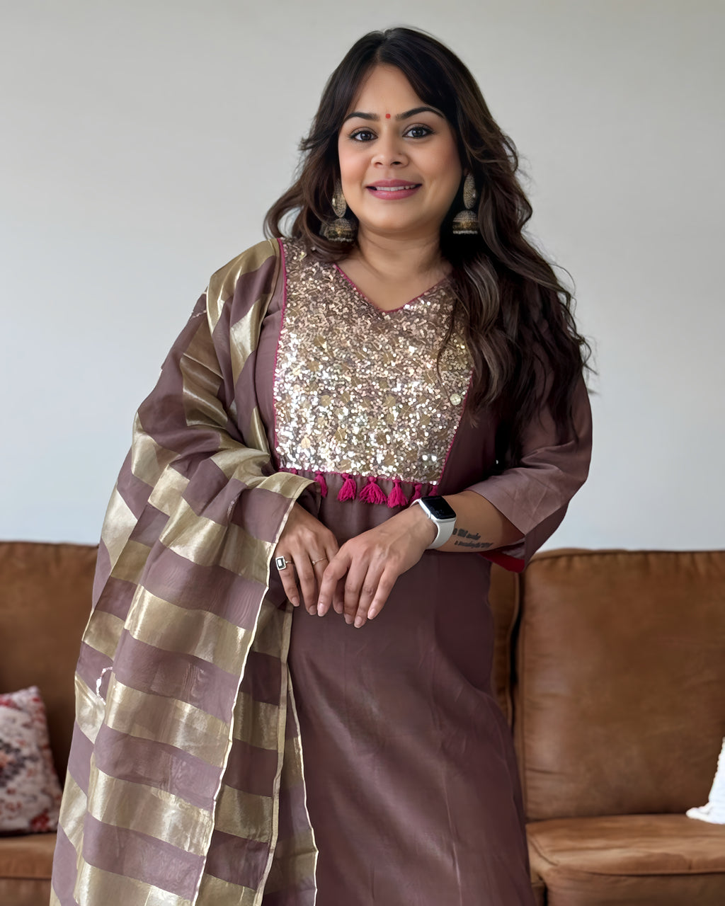 A woman wearing a brown and gold traditional outfit with a sequined chest panel and a striped dupatta, standing indoors in front of a brown sofa.