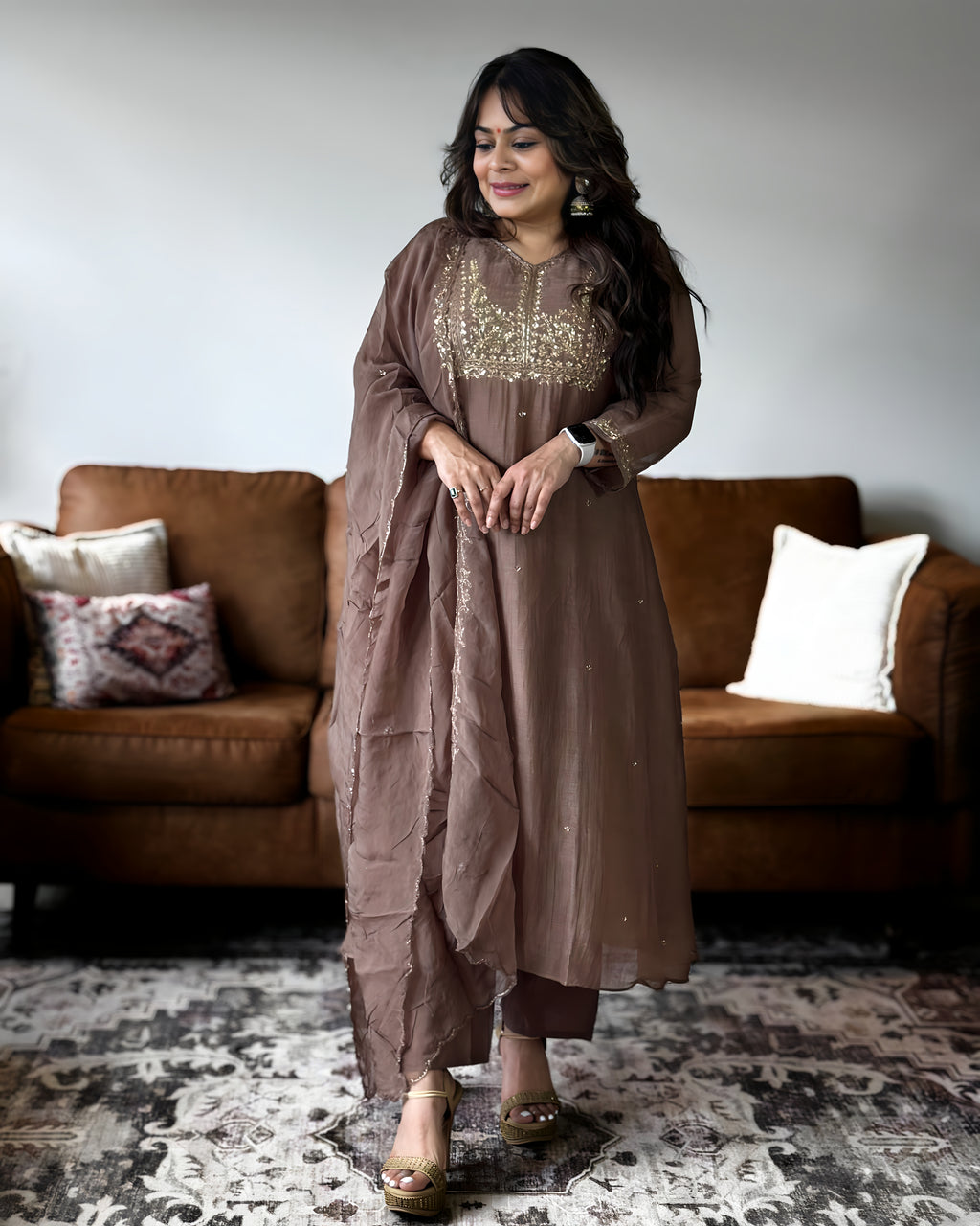 A woman models a brown traditional ethnic outfit with gold embroidery on the neckline, standing in front of a brown leather sofa in a living room setting.