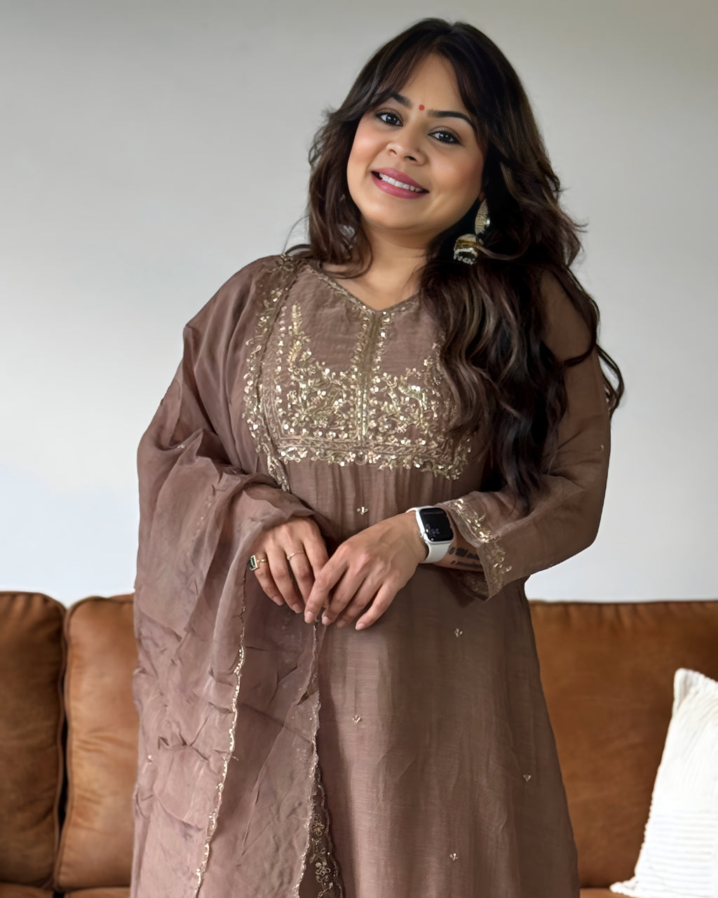 A woman wearing a brown traditional embroidered dress with a matching sheer dupatta, standing in front of a brown leather sofa.