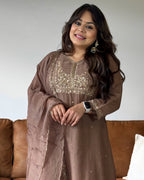 A woman wearing a brown traditional embroidered dress with a matching sheer dupatta, standing in front of a brown leather sofa.