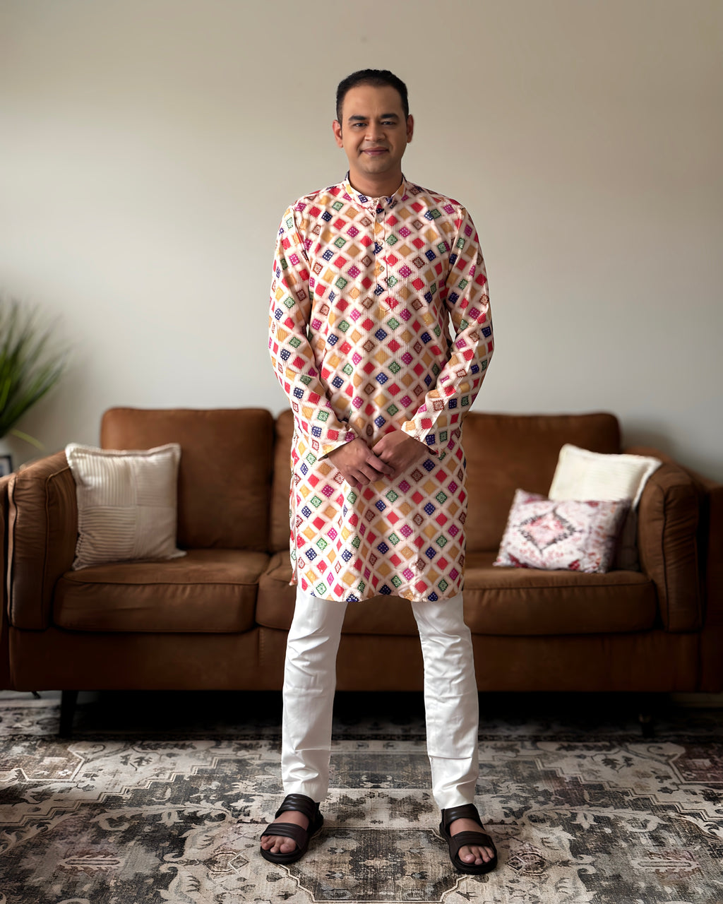 Man wearing a colorful patterned kurta standing in a living room with a brown sofa and patterned rug.