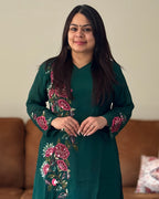 Woman wearing a dark green embroidered outfit standing in a room with a brown couch.