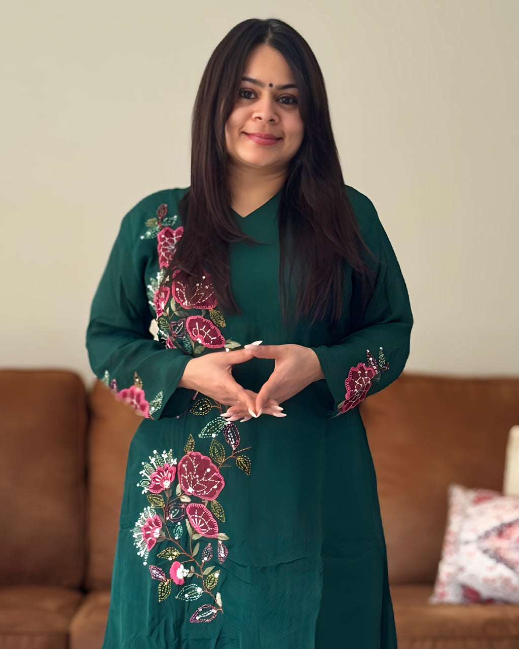 Woman wearing a green embroidered dress standing in a room with a brown couch and patterned pillow.