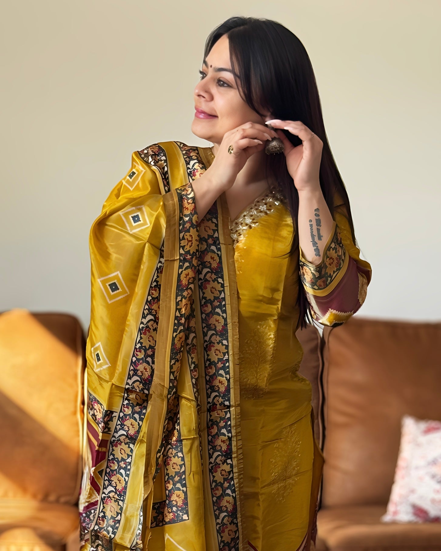 Woman wearing a yellow saree with a patterned border, sitting on a couch.