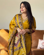 Woman wearing a traditional yellow saree with intricate patterns in a living room setting.