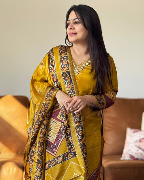 Woman wearing a traditional yellow saree with intricate patterns in a living room setting.