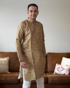 Man wearing a patterned kurta standing in a living room with a brown sofa and decorative pillows.