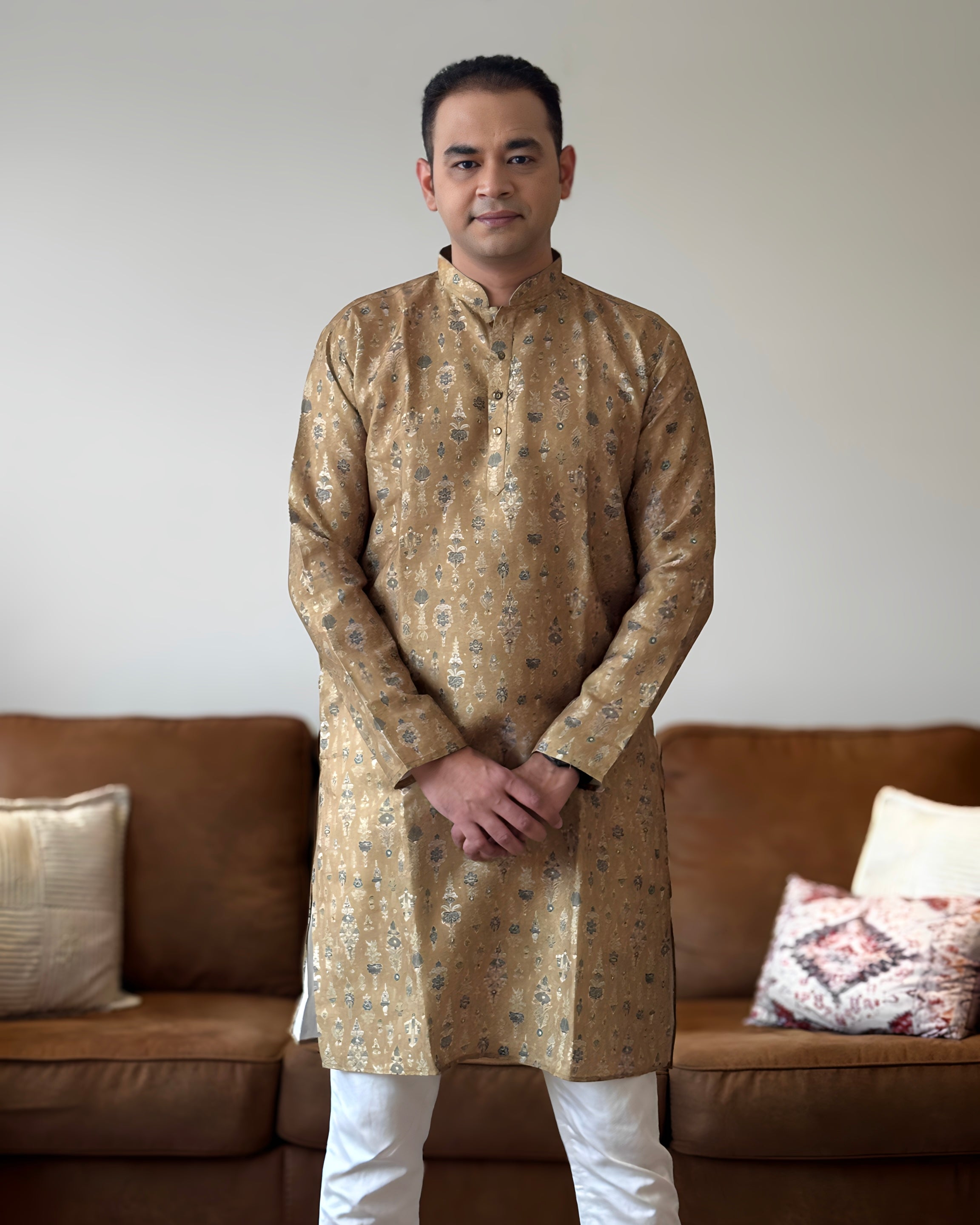Man wearing a patterned kurta standing in a living room with a brown sofa and decorative pillows.