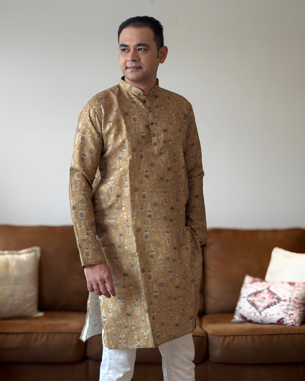 Man wearing a patterned kurta standing in a living room with a brown sofa and decorative pillows.