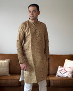 Man wearing a patterned kurta standing in a living room with a brown sofa and decorative pillows.