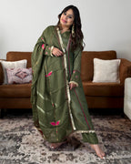 A woman models an olive green ethnic kurta set with pink floral embroidery and gold zari borders, standing in a living room setting with a brown sofa and patterned rug.