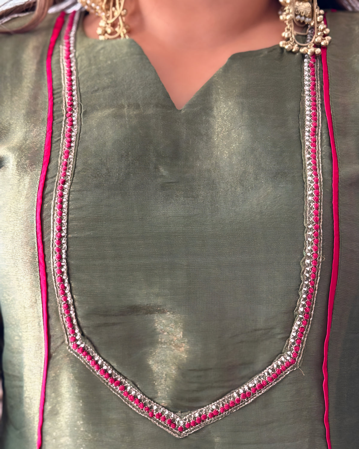 Close-up of a metallic olive green V-neck top with silver and pink embroidered neckline details, worn by a model with gold jewelry.