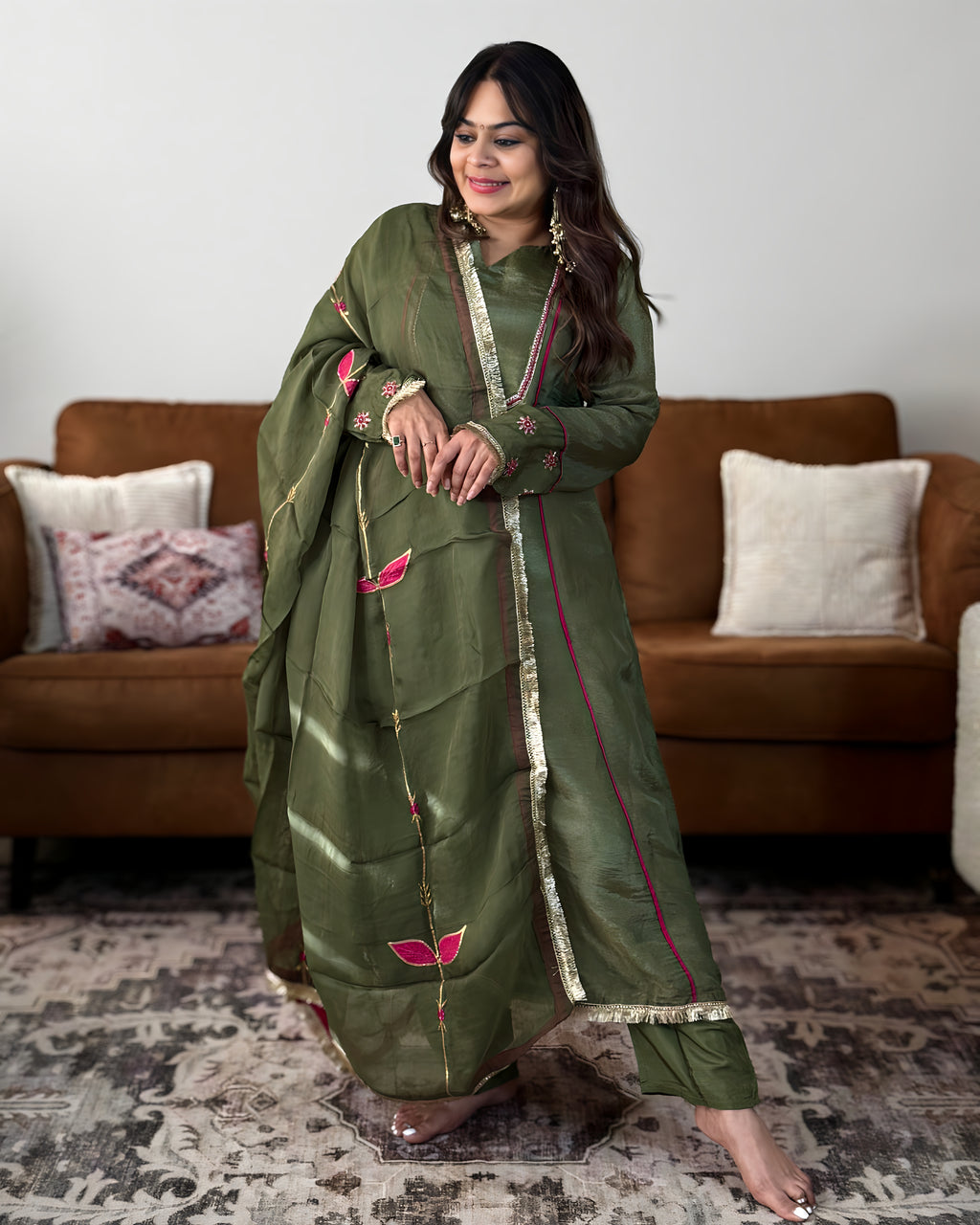 A woman models a traditional olive green salwar kameez set with pink floral embroidery and gold zari borders, standing in a living room setting with a brown sofa in the background.
