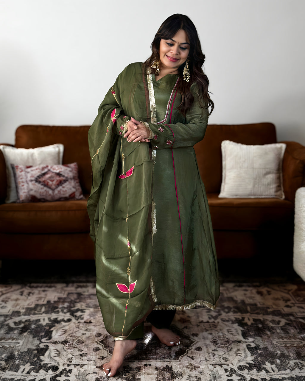 A woman modeling an olive green traditional outfit with pink and gold floral embroidery, standing in a living room setting with a brown sofa and patterned rug.