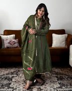 A woman modeling an olive green traditional outfit with pink and gold floral embroidery, standing in a living room setting with a brown sofa and patterned rug.