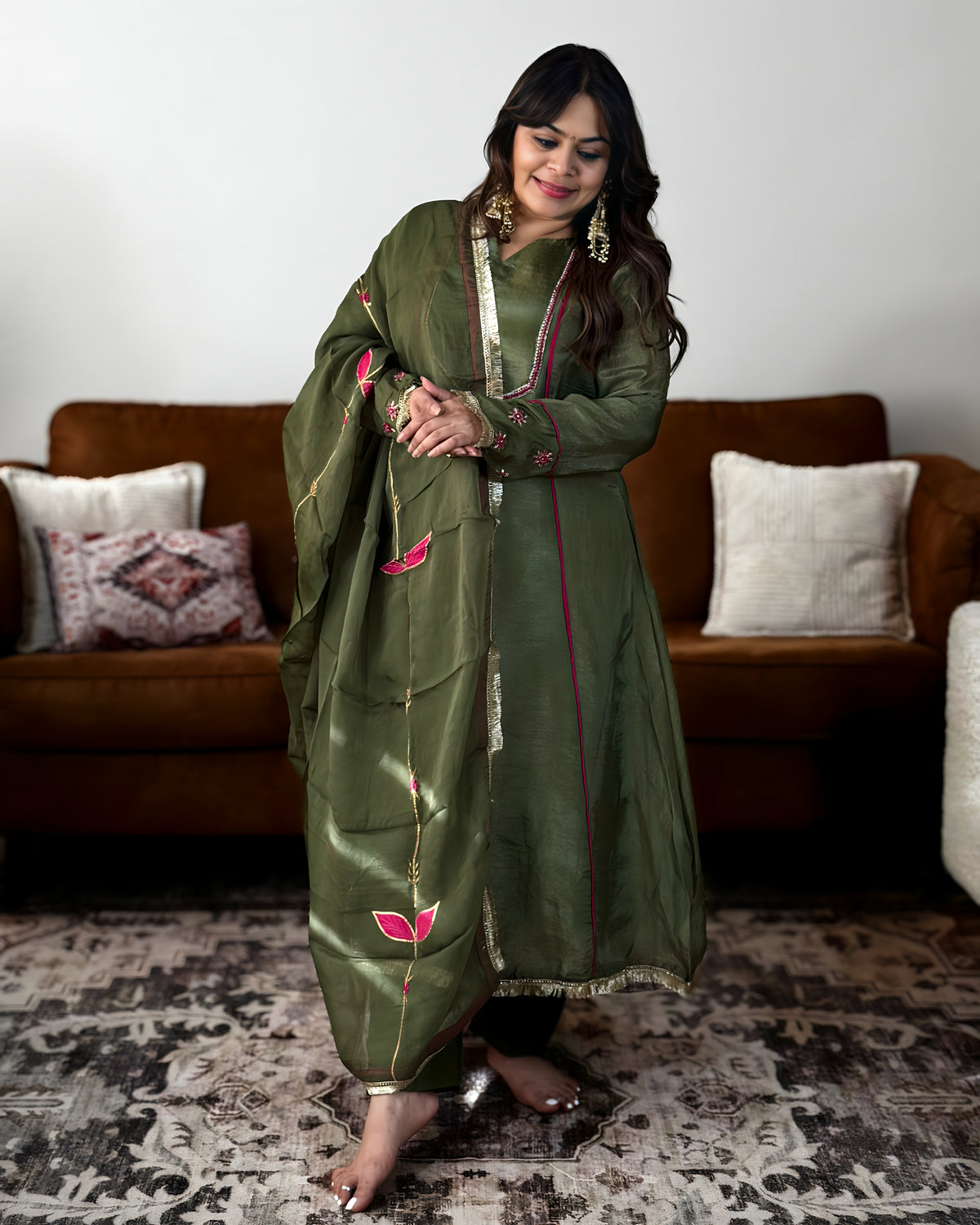 A woman modeling an olive green traditional outfit with pink and gold floral embroidery, standing in a living room setting with a brown sofa and patterned rug.