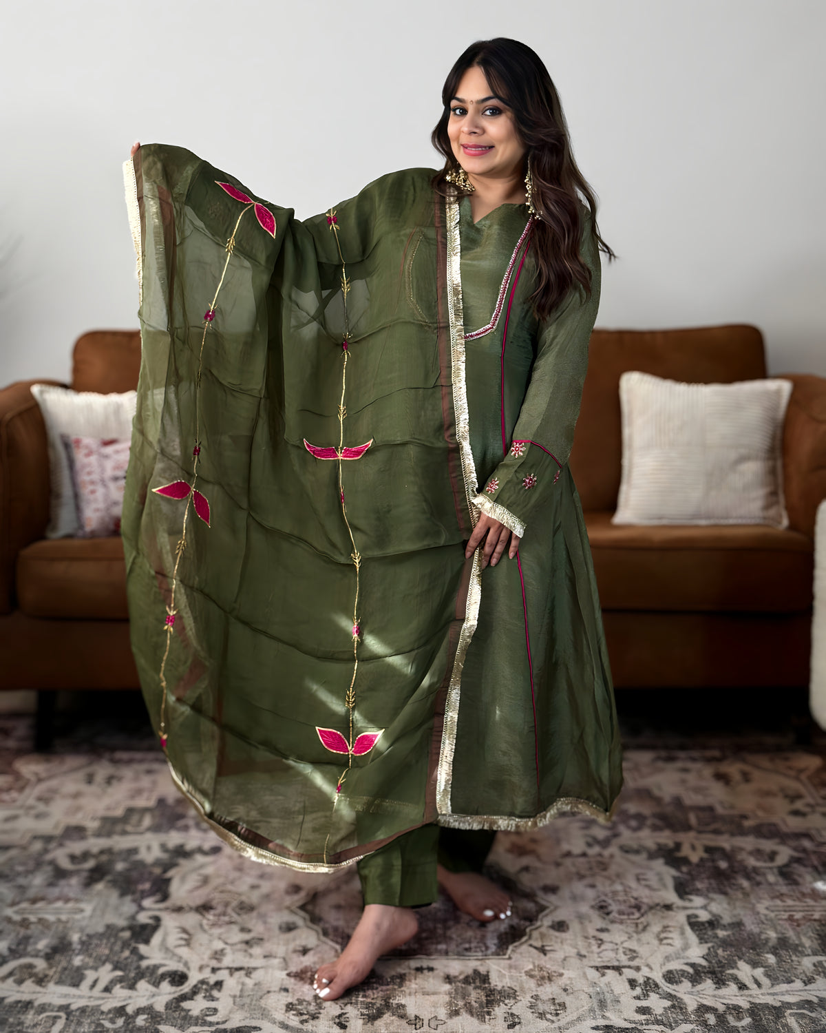 A woman models an olive green traditional Indian outfit with a matching dupatta, featuring pink floral embroidery and gold borders, set against a living room background with a brown sofa.