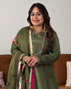 A woman wearing an olive green traditional outfit with gold and pink embroidered details, standing in front of a neutral wall with a brown sofa in the background.