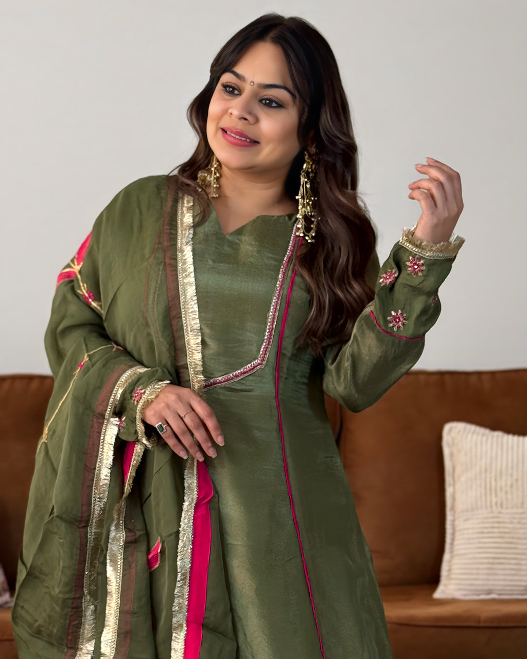 A woman wearing an olive green traditional South Asian salwar kameez suit with a matching dupatta, featuring gold zari borders and pink floral embroidery on the sleeves and neckline, posed in front of a brown leather sofa.