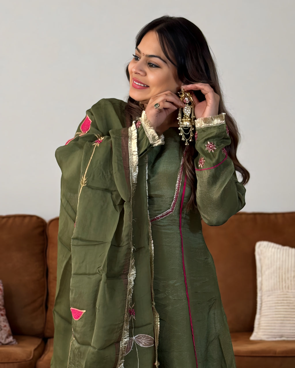 A woman wearing an olive green traditional outfit with pink and gold embroidery, holding a matching embroidered shawl, standing in front of a brown sofa.