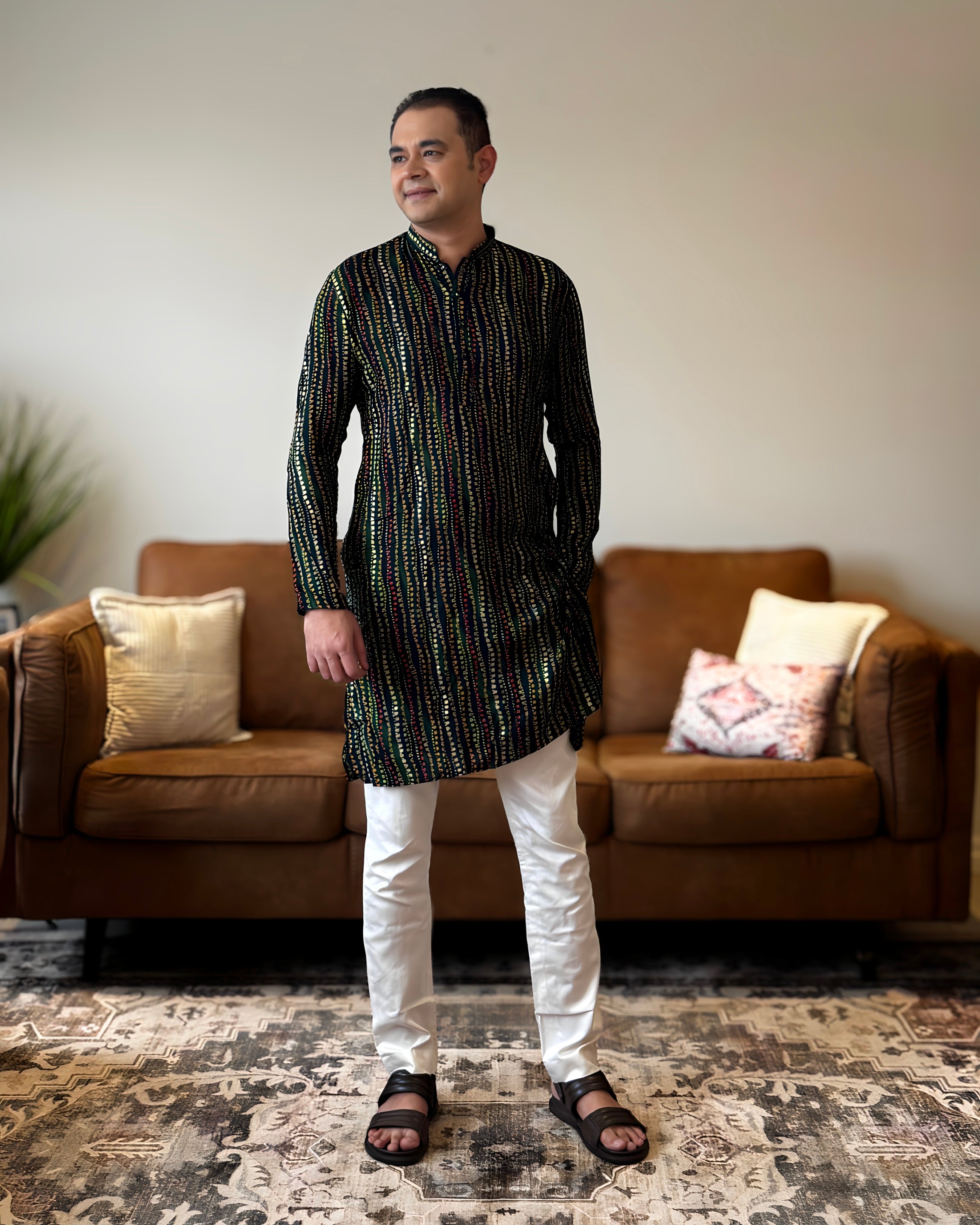 Man wearing a black and white patterned kurta with white pants standing in a living room.
