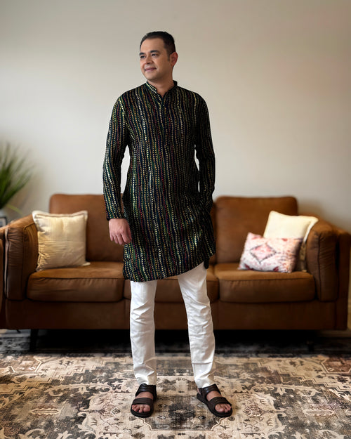 Man wearing a black and white patterned kurta with white pants standing in a living room.