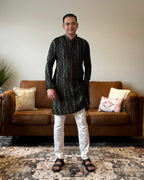 Man wearing a black patterned kurta with white pants standing in a living room.