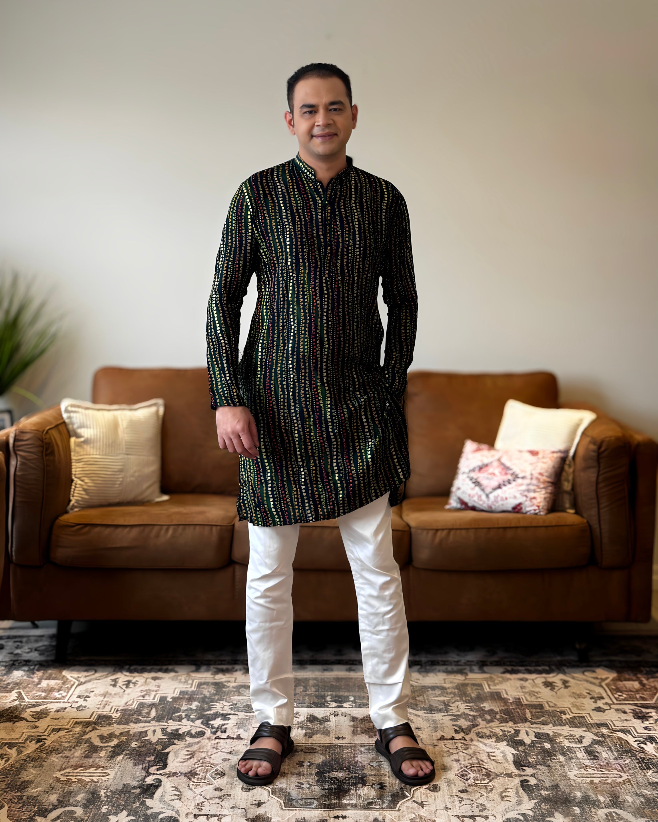 Man wearing a black patterned kurta with white pants standing in a living room.