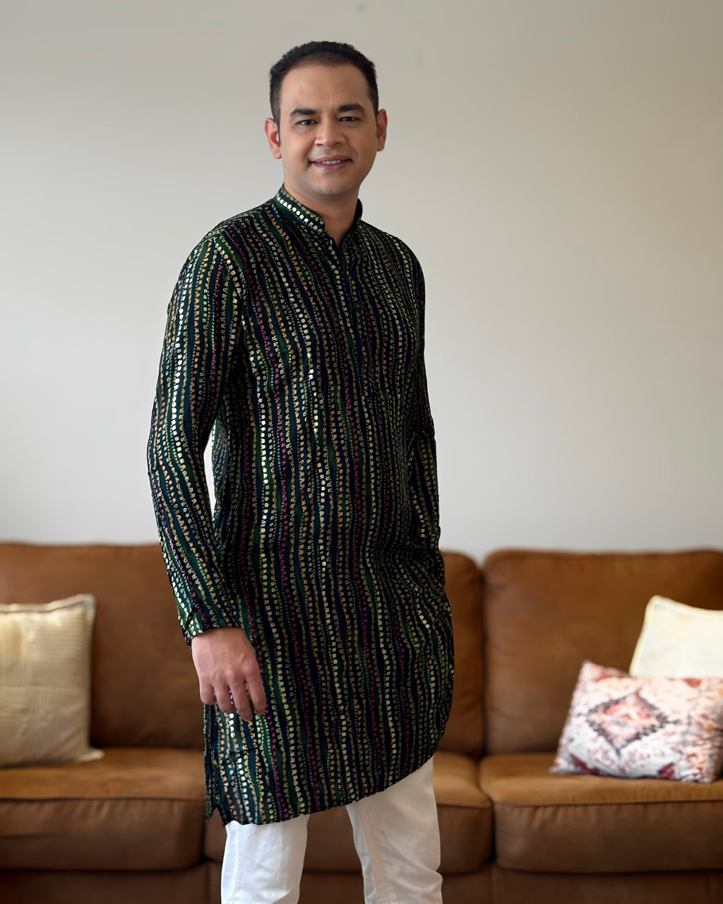 Man wearing a patterned kurta standing in a living room with a brown sofa and decorative pillows.
