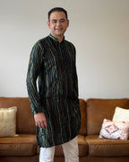 Man wearing a patterned kurta standing in a living room with a brown sofa and decorative pillows.