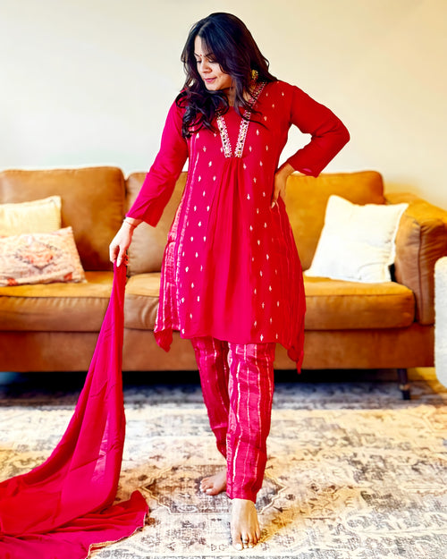 Woman in a red traditional outfit standing in a living room with a couch and pillows.