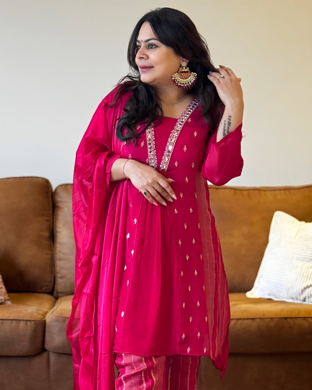 Woman in a bright pink traditional outfit with white patterns, standing in a room with a brown couch and white wall.