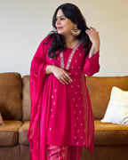 Woman in a bright pink traditional outfit with white patterns, standing in a room with a brown couch and white wall.