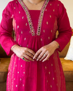 Person wearing a bright pink traditional outfit with intricate designs, sitting on a brown couch.