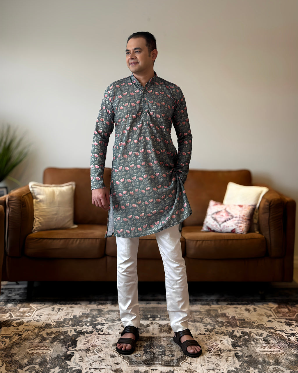 Man wearing a patterned kurta standing in a living room with a brown sofa and decorative pillows.