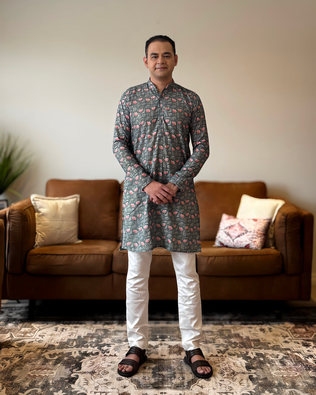 Man wearing a patterned kurta standing in a living room with a brown sofa and decorative pillows.
