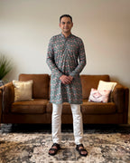 Man wearing a patterned kurta standing in a living room with a brown sofa and decorative pillows.