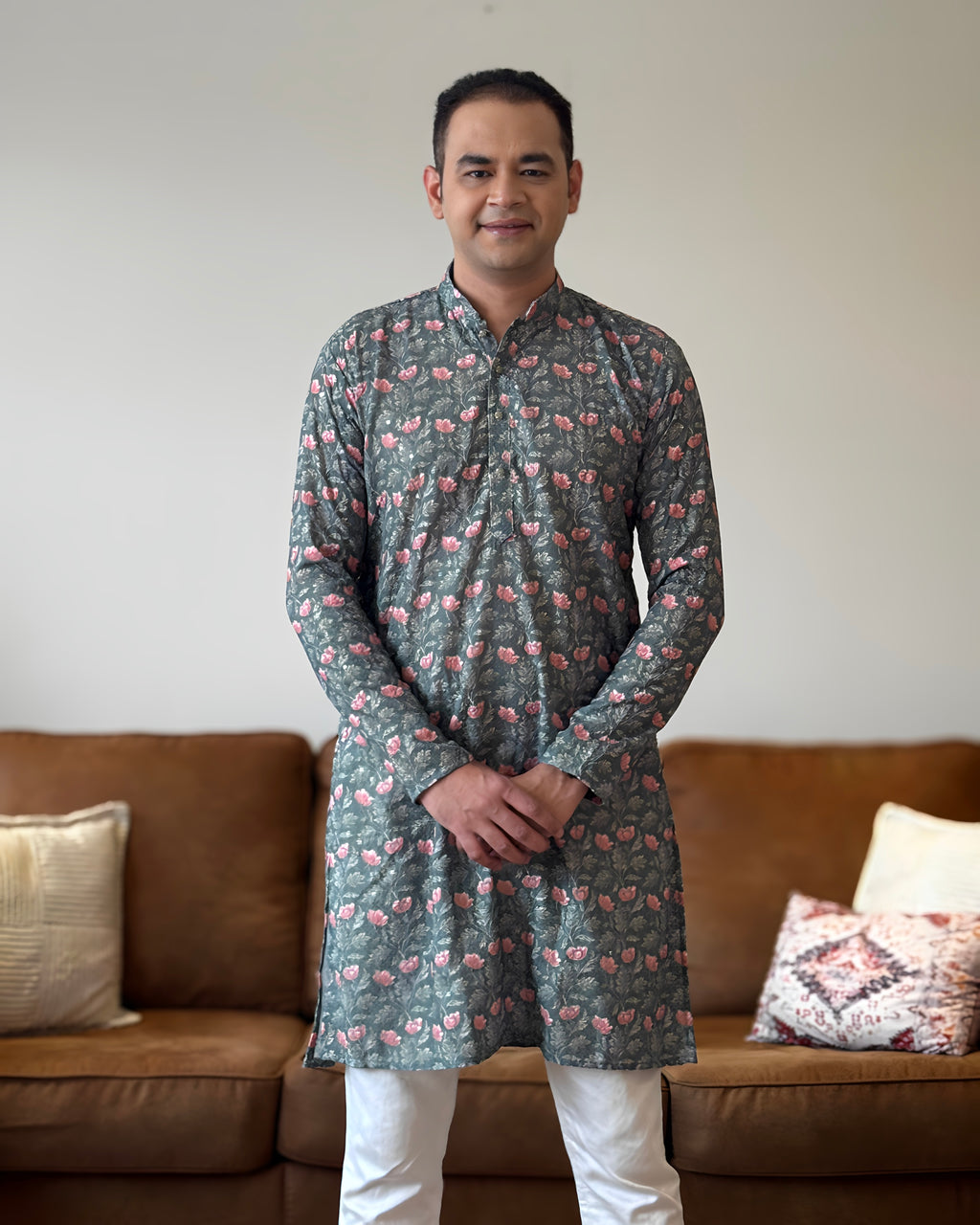 Man wearing a patterned kurta standing in a living room with a brown sofa and decorative pillows.