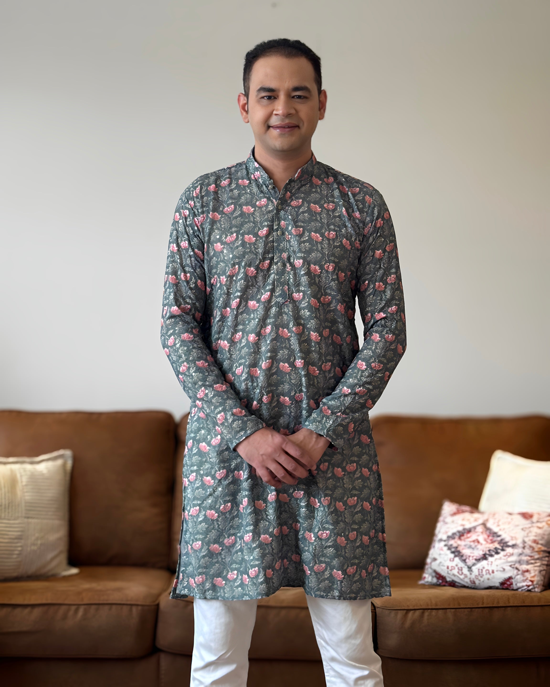 Man wearing a patterned kurta standing in a living room with a brown sofa and decorative pillows.