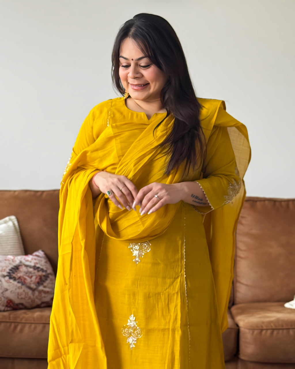 Woman in a yellow saree with a brown couch and decorative pillows in the background