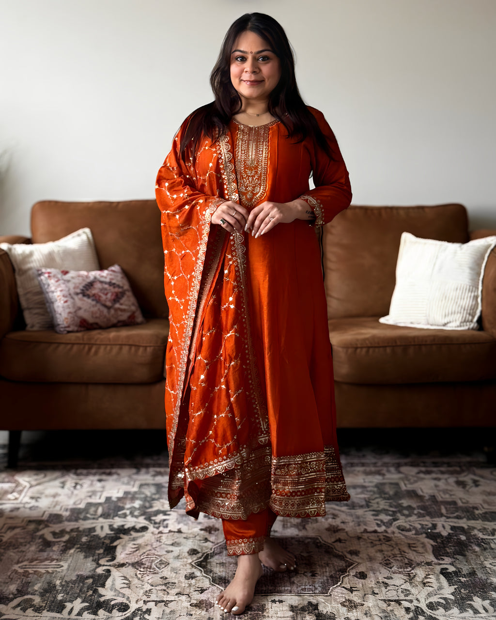 Woman in an orange traditional outfit standing in a living room with a brown sofa and patterned rug.