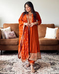 Woman in an orange traditional outfit standing in a living room with a brown sofa and patterned rug.